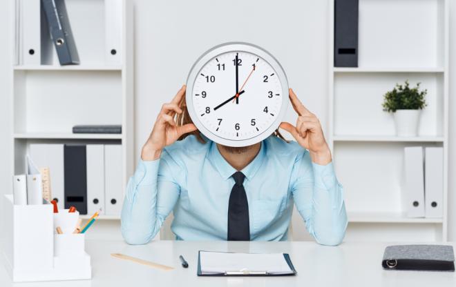 Une personne assise derrière son bureau tient une horloge devant son visage.